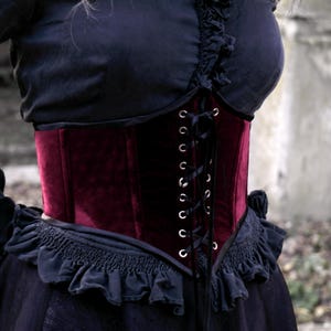 May include: A burgundy velvet corset with a lace-up front and silver eyelets. The corset is worn over a black top with ruffled detailing. The overall style is gothic and elegant.