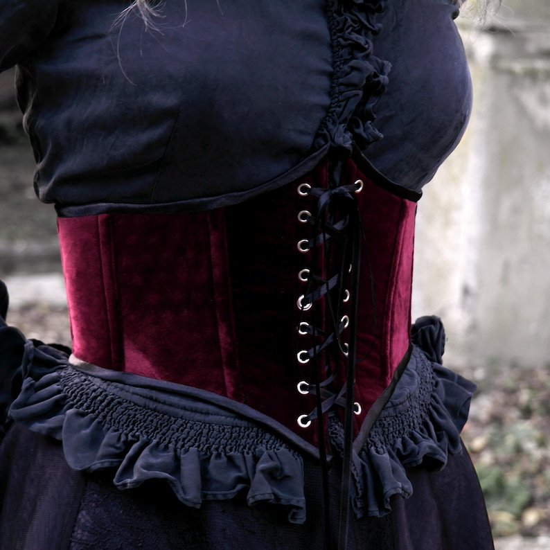 May include: A burgundy velvet corset with a lace-up front and silver eyelets. The corset is worn over a black top with ruffled detailing. The overall style is gothic and elegant.