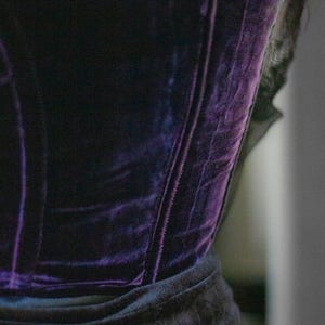 May include: A close-up of a dark purple velvet corset. The corset has a structured design with vertical seams and a black trim. The fabric has a rich, textured appearance, and the overall style suggests a gothic or vintage aesthetic.