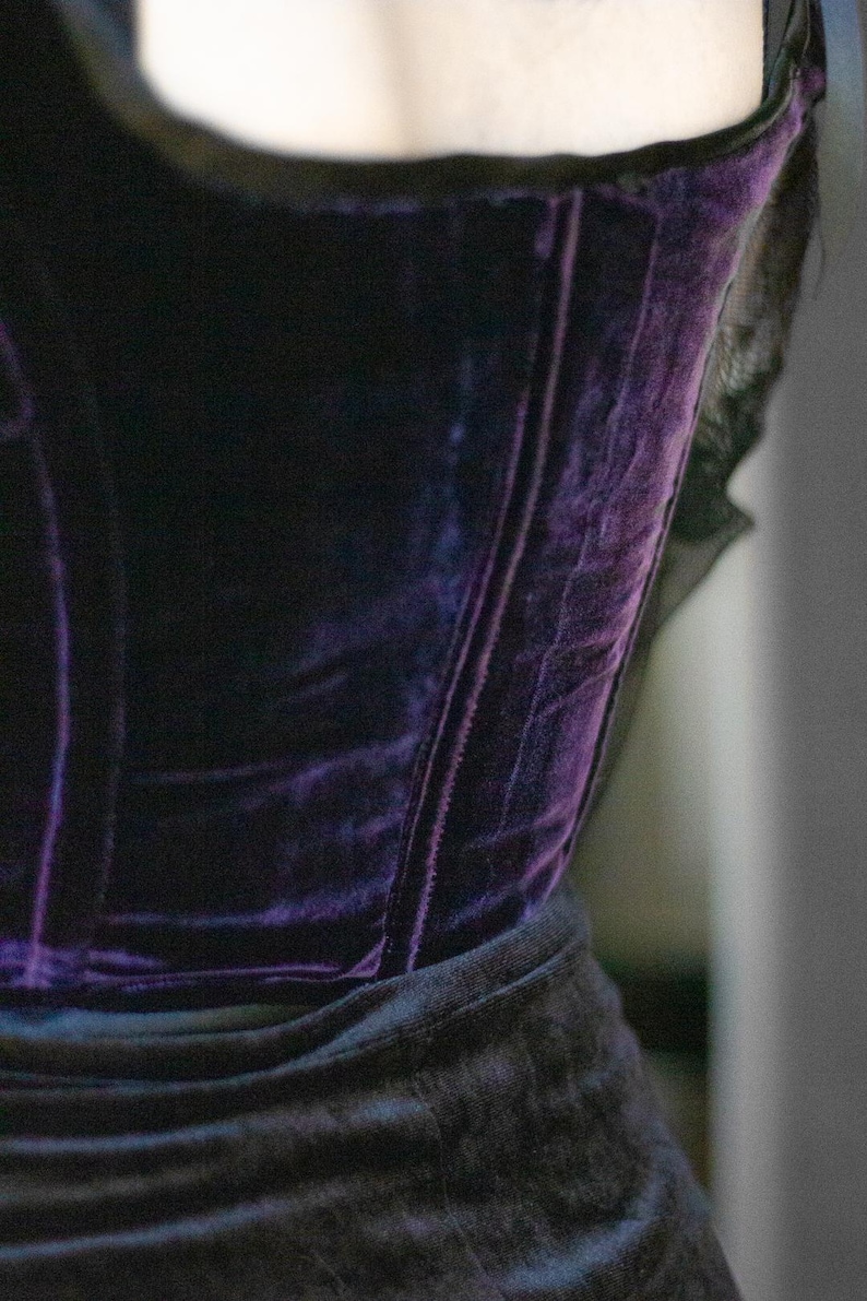 May include: A close-up of a dark purple velvet corset. The corset has a structured design with vertical seams and a black trim. The fabric has a rich, textured appearance, and the overall style suggests a gothic or vintage aesthetic.
