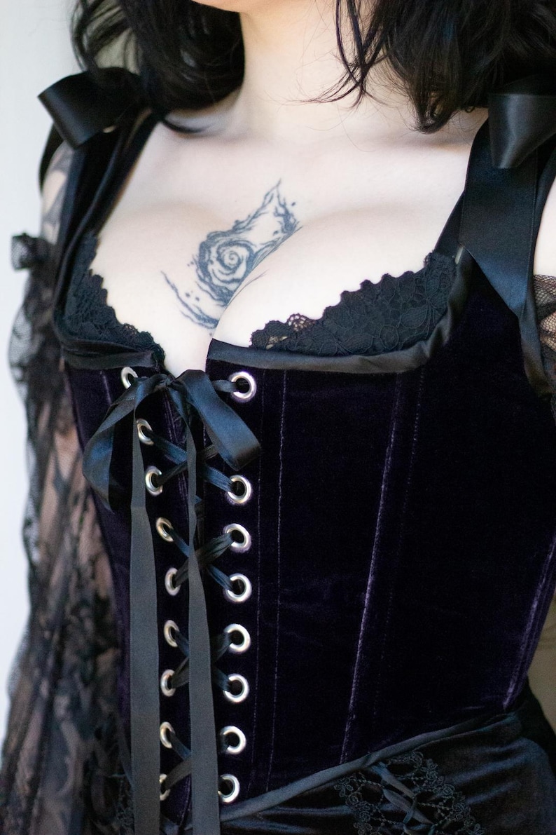 May include: Close-up of a black velvet corset with a lace trim. The corset features a lace-up front with silver eyelets and black satin ribbon. The straps are adorned with black bows. The overall style is gothic.
