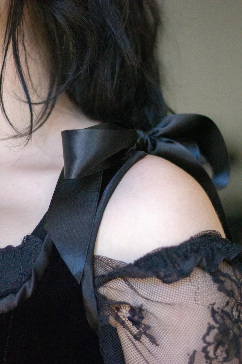 May include: Close-up of a black top with a satin bow detail on the shoulder. The top features a lace sleeve with a floral pattern. The overall style suggests a gothic or romantic aesthetic, suitable for special occasions.