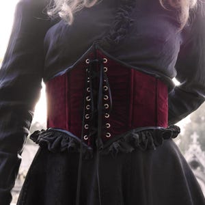May include: A burgundy velvet corset with black lacing and eyelets. The corset is worn over a black top and dress, creating a layered effect. The design features a ruffled trim at the top and bottom.