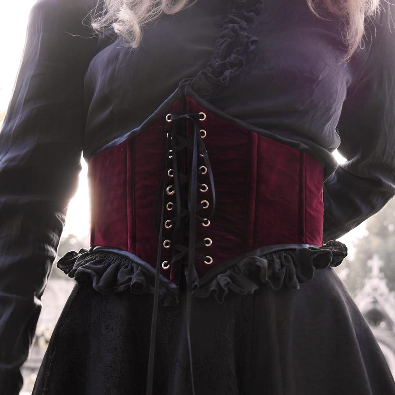 May include: A burgundy velvet corset with black lacing and eyelets. The corset is worn over a black top and dress, creating a layered effect. The design features a ruffled trim at the top and bottom.