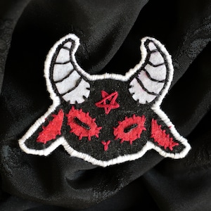 Może przedstawiać: Czarna łatka z filcu z białymi szwami, przedstawiająca kreskówkowego diabła z czerwonymi oczami i rogami. Diabeł ma czerwony pentagram na czole.