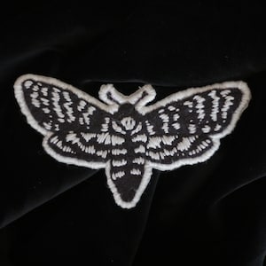 Peut inclure: Un patch brodé de papillon de nuit noir et blanc avec un design détaillé. Le papillon de nuit a un motif d'œil grand et proéminent sur ses ailes.
