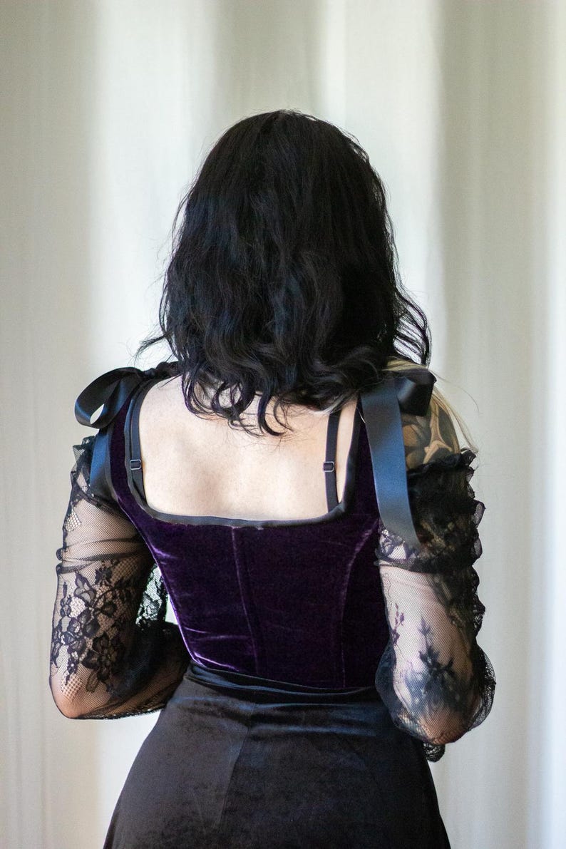 May include: A dark purple velvet corset top with black lace sleeves and black ribbon shoulder ties. The top has a square neckline and is paired with a black skirt. The overall style is gothic and elegant.