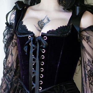 May include: A black velvet corset with a deep purple hue, featuring a lace-up front and delicate lace trim. The corset has black ribbon straps and is paired with sheer, lacy sleeves. The design is gothic and elegant.