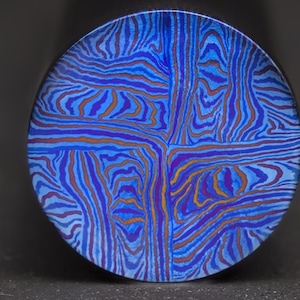 Puede incluir: Un disco redondo de metal azul y morado con un patrón de rayas en espiral. El patrón está formado por líneas finas y onduladas en tonos azules, morados y naranjas.