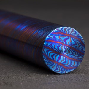 Puede incluir: Primer plano de una varilla metálica redonda de color azul, morado y naranja con un patrón de espiral. La varilla está sobre una superficie gris.