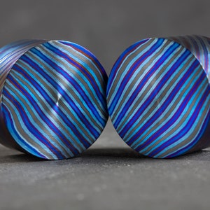 Puede incluir: Dos piezas redondas y cilíndricas de metal con un patrón de rayas azules, moradas y grises. El metal tiene un acabado pulido y brillante.