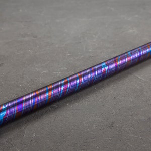 Puede incluir: Una varilla metálica larga y delgada con un patrón de espiral colorido en tonos azules, morados y naranjas. La varilla está sobre una superficie gris.