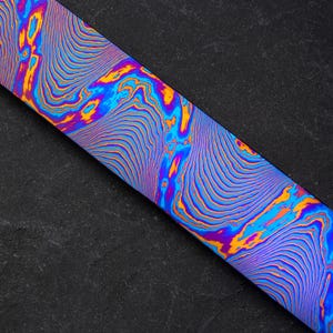 Pode incluir: Close-up de uma superfície metálica com padrões azuis, roxos e laranja. A superfície tem um design ondulado e abstrato.