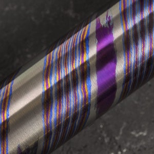 Puede incluir: Primer plano de una varilla metálica con un patrón de rayas multicolores. El patrón se crea mediante un proceso llamado anodizado, que crea una fina capa de óxido en la superficie del metal. La capa de óxido se tiñe luego de diferentes colores para crear el patrón.
