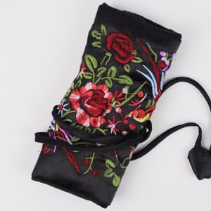 May include: Black satin roll-up case with vibrant floral embroidery in red, green, and yellow. The case has a drawstring closure and is designed to hold items. The embroidered flowers and leaves create a decorative pattern.