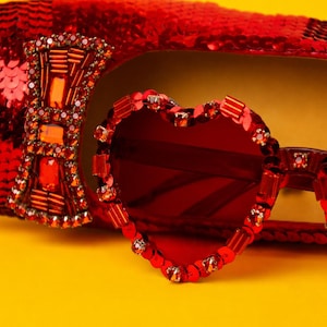 May include: A red sequined shoe featuring a large, rectangular, crystal-embellished bow ornament, positioned next to heart-shaped sunglasses with red frames decorated with matching red crystals and sequins, all set against a bright yellow background.