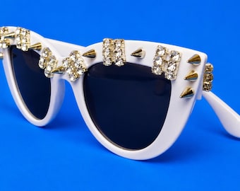 Puede incluir: Gafas de sol blancas estilo ojo de gato con lentes oscuras y una montura adornada con púas metálicas doradas y grupos de brillantes transparentes a lo largo del borde superior y los bordes exteriores, creando una textura llamativa.
