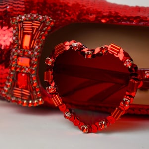May include: A close-up of a red sequined shoe with a decorative bow and a pair of heart-shaped sunglasses. The sunglasses and shoe are embellished with red rhinestones.