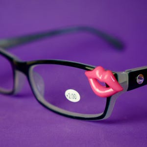 May include: Black framed reading glasses with pink lips on the arms and a +2.00 label on the right lens.