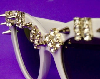 Puede incluir: Gafas de sol blancas con pedrería y pinchos plateados. Las gafas tienen una montura y lentes blancas.