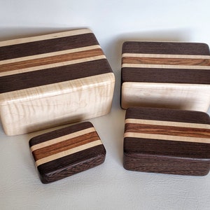 May include: Four wooden boxes of varying sizes. Each box is rectangular with a striped design. The stripes alternate between dark brown, light brown, and natural wood tones. The boxes are arranged on a light-colored surface.