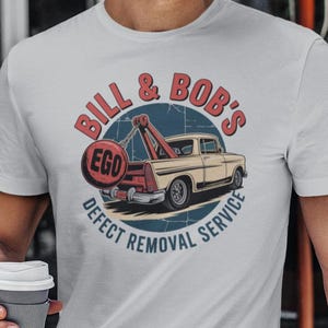May include: White t-shirt with a vintage-style graphic. The design features a tow truck and the text "Bill & Bob's Defect Removal Service" in a circular emblem. The shirt is worn by a person.