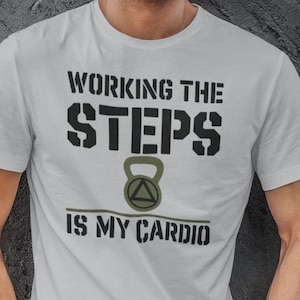 May include: A light gray t-shirt with the text "WORKING THE STEPS IS MY CARDIO" in black. The shirt features a dumbbell graphic. The person in the image is wearing blue jeans and has their hands in their pockets.