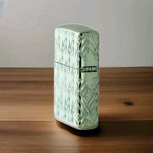 K&ouml;nnte beinhalten: Ein metallisch gr&uuml;nes Feuerzeug mit einem gepr&auml;gten Blattmuster. Das rechteckige Feuerzeug hat abgerundete Kanten und einen Klappdeckel. Das Design erzeugt eine strukturierte Oberfl&auml;che. Das Feuerzeug steht vor einem Holzgrund.