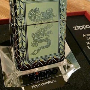 K&ouml;nnte beinhalten: Ein Zippo-Feuerzeug mit einer detaillierten Drachengravur auf der Vorderseite. Das Feuerzeug hat eine silberne und goldene Oberfl&auml;che mit dekorativen Mustern an den R&auml;ndern. Das Zippo-Logo ist auf der Verpackung sichtbar.