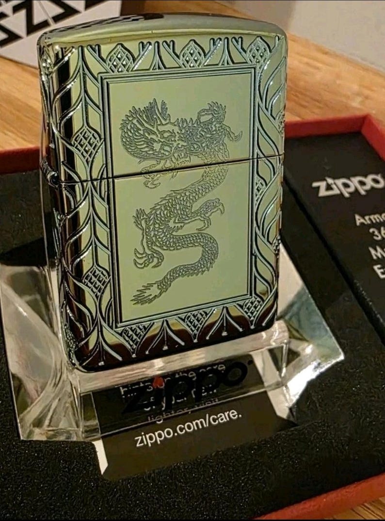 K&ouml;nnte beinhalten: Ein Zippo-Feuerzeug mit einer detaillierten Drachengravur auf der Vorderseite. Das Feuerzeug hat eine silberne und goldene Oberfl&auml;che mit dekorativen Mustern an den R&auml;ndern. Das Zippo-Logo ist auf der Verpackung sichtbar.