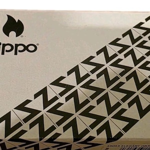 K&ouml;nnte beinhalten: Rechteckige Schachtel mit dem Zippo-Logo und einem schwarzen Flammen-Symbol. Die Schachtel hat ein diagonales Muster aus schwarzen Zickzacklinien auf hellem Hintergrund. Der Text "Zippo" ist schwarz gedruckt.