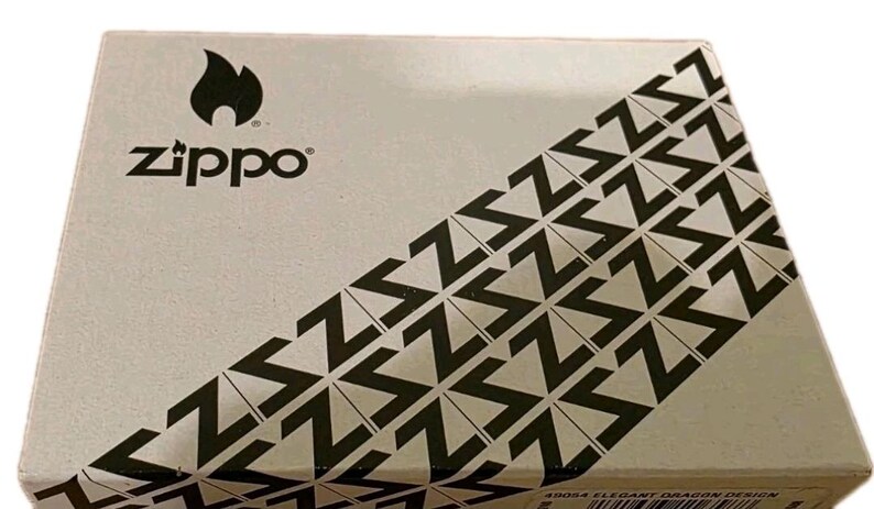 K&ouml;nnte beinhalten: Rechteckige Schachtel mit dem Zippo-Logo und einem schwarzen Flammen-Symbol. Die Schachtel hat ein diagonales Muster aus schwarzen Zickzacklinien auf hellem Hintergrund. Der Text "Zippo" ist schwarz gedruckt.