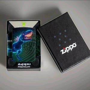 Puede incluir: Un encendedor Zippo con un dise&ntilde;o de Tyrannosaurus Rex azul ne&oacute;n sobre un fondo de rejilla verde. El encendedor est&aacute; en su caja negra, con el logotipo de Zippo en blanco. La caja est&aacute; abierta, revelando el encendedor y su embalaje.