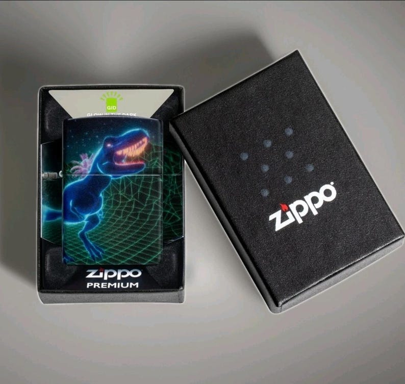 Puede incluir: Un encendedor Zippo con un dise&ntilde;o de Tyrannosaurus Rex azul ne&oacute;n sobre un fondo de rejilla verde. El encendedor est&aacute; en su caja negra, con el logotipo de Zippo en blanco. La caja est&aacute; abierta, revelando el encendedor y su embalaje.