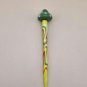 May include: A whimsical mushroom-shaped decorative pin. The top is a dark green with white polka dots. The stem is yellow with red, blue, and yellow swirls. The pin is a unique accessory.