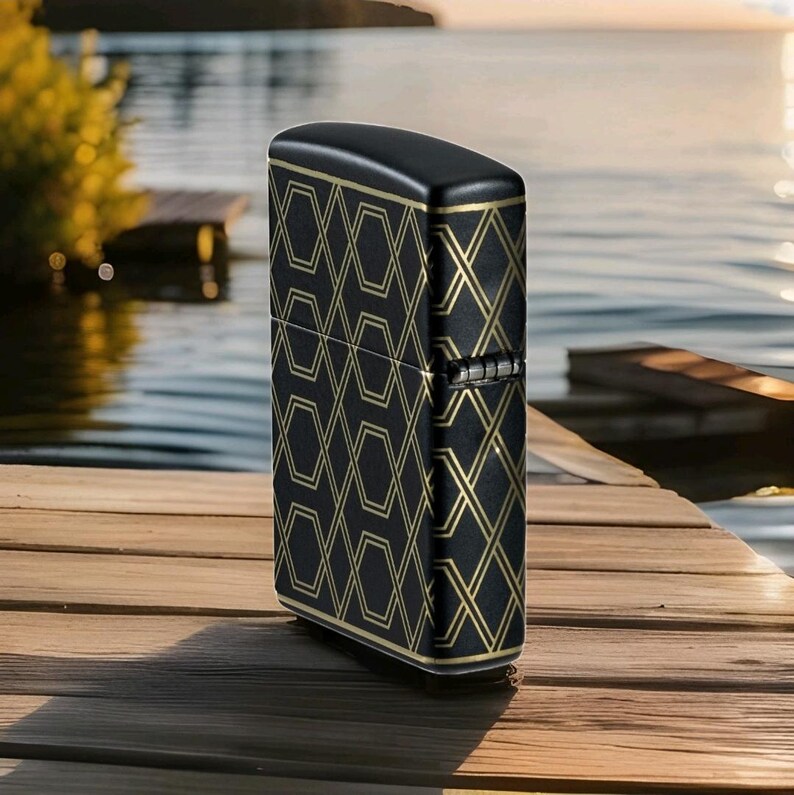 Puede incluir: Encendedor negro con un patr&oacute;n geom&eacute;trico dorado. El encendedor tiene forma rectangular con bordes redondeados. El patr&oacute;n dorado es un dise&ntilde;o repetitivo de formas conectadas. El encendedor est&aacute; sobre una superficie de madera cerca del agua.