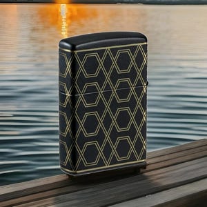 Puede incluir: Encendedor negro con un patr&oacute;n geom&eacute;trico dorado. El encendedor tiene forma rectangular con bordes redondeados y una tapa con bisagras. El patr&oacute;n dorado es un dise&ntilde;o repetitivo de hex&aacute;gonos y l&iacute;neas entrelazadas. El encendedor est&aacute; sobre una superficie de madera.