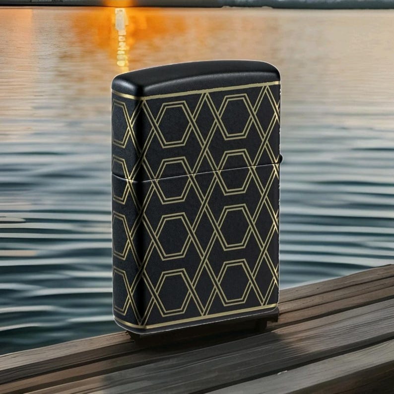 Puede incluir: Encendedor negro con un patr&oacute;n geom&eacute;trico dorado. El encendedor tiene forma rectangular con bordes redondeados y una tapa con bisagras. El patr&oacute;n dorado es un dise&ntilde;o repetitivo de hex&aacute;gonos y l&iacute;neas entrelazadas. El encendedor est&aacute; sobre una superficie de madera.