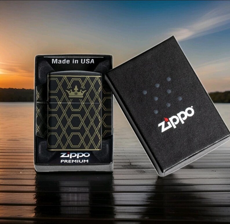 Puede incluir: Encendedor Zippo negro con un patr&oacute;n geom&eacute;trico dorado y dise&ntilde;o de corona, exhibido en su caja negra original. La caja est&aacute; abierta, revelando el encendedor y las palabras "Made in USA". El logotipo de Zippo es visible en la caja.