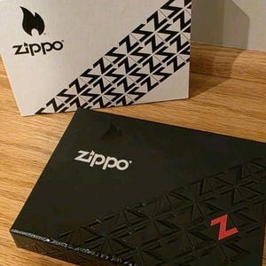 K&ouml;nnte beinhalten: Zwei Zippo-Boxen werden gezeigt. Eine ist wei&szlig; mit einem schwarzen Flammenlogo und diagonalen schwarzen Z-Mustern. Die andere ist schwarz mit dem Zippo-Logo und einem strukturierten Muster mit einem roten Z.