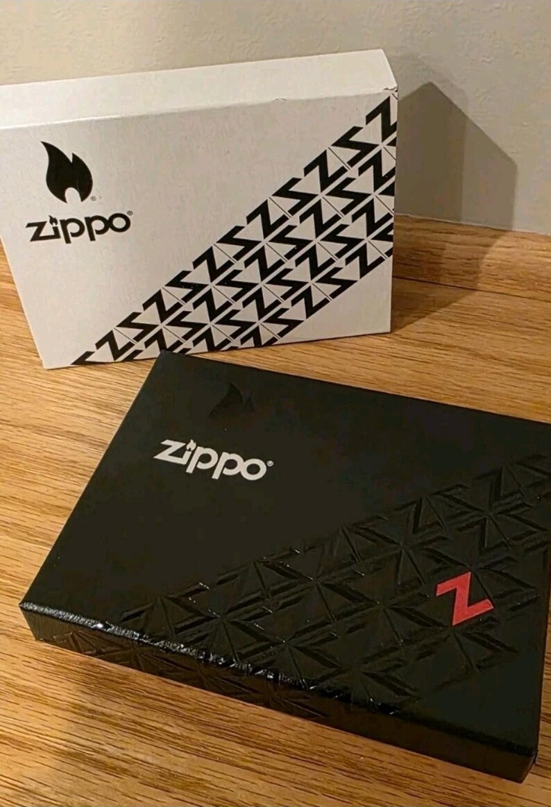K&ouml;nnte beinhalten: Zwei Zippo-Boxen werden gezeigt. Eine ist wei&szlig; mit einem schwarzen Flammenlogo und diagonalen schwarzen Z-Mustern. Die andere ist schwarz mit dem Zippo-Logo und einem strukturierten Muster mit einem roten Z.