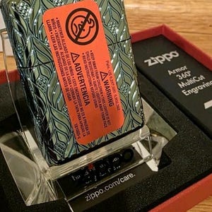 K&ouml;nnte beinhalten: Ein Zippo-Feuerzeug in Gr&uuml;n und Silber mit einem Bl&auml;ttermuster, in der Originalverpackung. Die Verpackung enth&auml;lt eine rote Schachtel und einen transparenten Kunststoffst&auml;nder. Das Warnetikett ist deutlich sichtbar.