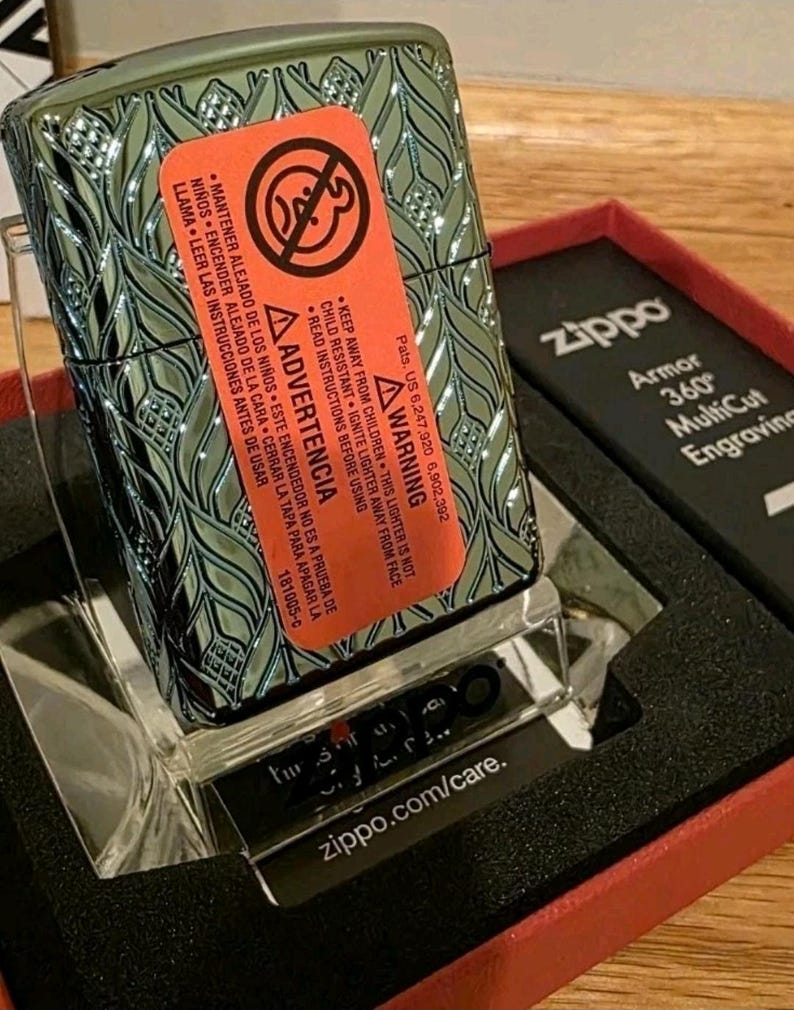 K&ouml;nnte beinhalten: Ein Zippo-Feuerzeug in Gr&uuml;n und Silber mit einem Bl&auml;ttermuster, in der Originalverpackung. Die Verpackung enth&auml;lt eine rote Schachtel und einen transparenten Kunststoffst&auml;nder. Das Warnetikett ist deutlich sichtbar.