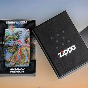 Może przedstawiać: Zippo Premium zapalniczka ze wzorem smoka w pudełku prezentowym. Zapalniczka posiada kolorową ilustrację smoka na jasnoniebieskim tle. Pudełko jest czarne z białym logo Zippo i napisem "Made in USA".