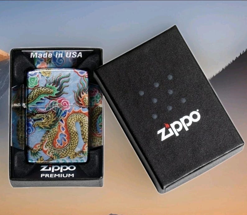 Może przedstawiać: Zippo Premium zapalniczka ze wzorem smoka w pudełku prezentowym. Zapalniczka posiada kolorową ilustrację smoka na jasnoniebieskim tle. Pudełko jest czarne z białym logo Zippo i napisem "Made in USA".