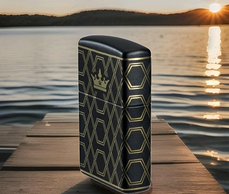 Puede incluir: Encendedor negro con un patr&oacute;n geom&eacute;trico dorado y un emblema de corona. El encendedor est&aacute; sobre un muelle de madera, con un lago y una puesta de sol de fondo. El encendedor tiene forma rectangular.