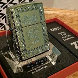 K&ouml;nnte beinhalten: Ein gr&uuml;nes Zippo-Feuerzeug mit Drachengravur, pr&auml;sentiert in einer roten Schachtel. Das Feuerzeug hat einen dekorativen Rand und den Text "Zippo Armor 360&deg; MultiCut Engraving". Es steht auf einem transparenten St&auml;nder.