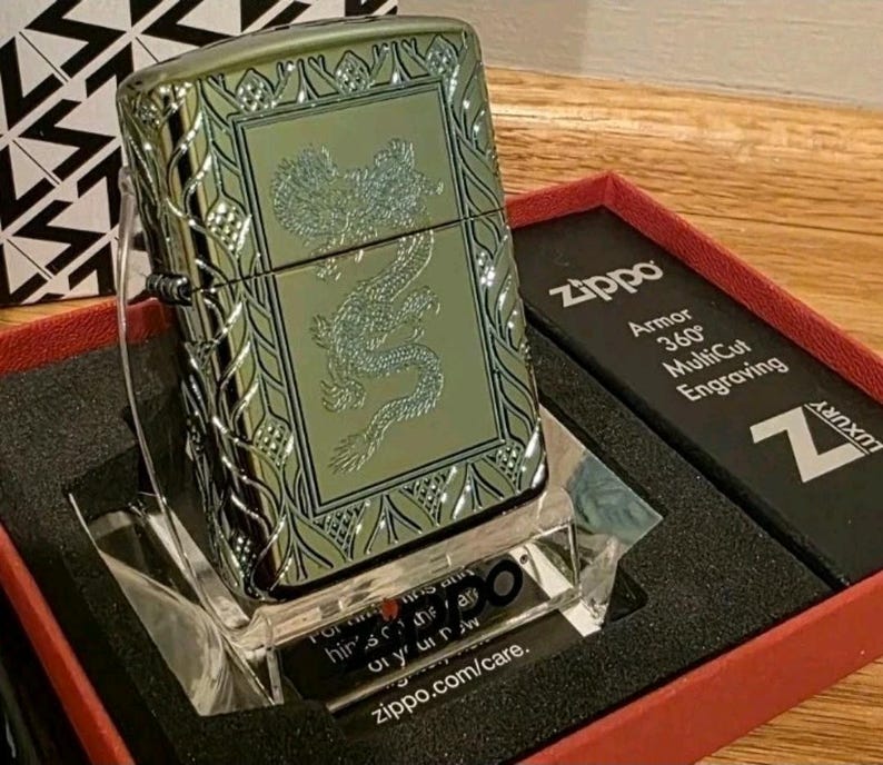 K&ouml;nnte beinhalten: Ein gr&uuml;nes Zippo-Feuerzeug mit Drachengravur, pr&auml;sentiert in einer roten Schachtel. Das Feuerzeug hat einen dekorativen Rand und den Text "Zippo Armor 360&deg; MultiCut Engraving". Es steht auf einem transparenten St&auml;nder.