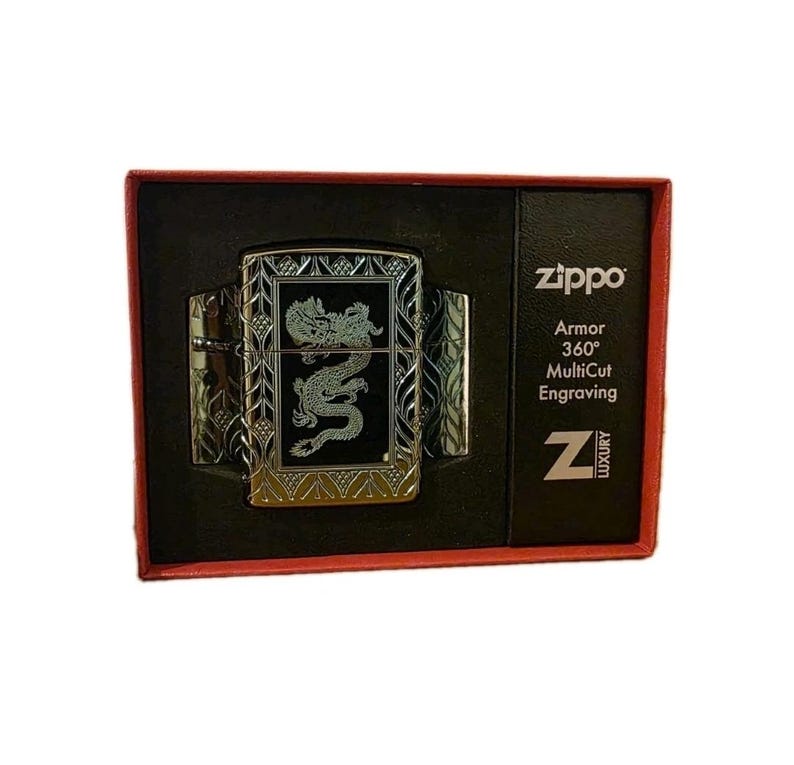 K&ouml;nnte beinhalten: Ein silberfarbenes Zippo-Feuerzeug mit Drachengravur, pr&auml;sentiert in einer schwarzen Box mit rotem Rand. Das Feuerzeug hat aufwendige Details und das Zippo-Logo, zusammen mit dem Text "Armor 360&deg; MultiCut Engraving".