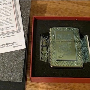 K&ouml;nnte beinhalten: Ein silbernes Zippo-Feuerzeug mit eingraviertem Design, pr&auml;sentiert in einer rot-schwarzen Box. Die Box enth&auml;lt eine Garantie und den Text "Armor 360&deg; MultiCut Engraving". Das Feuerzeug ist ein Luxusartikel.